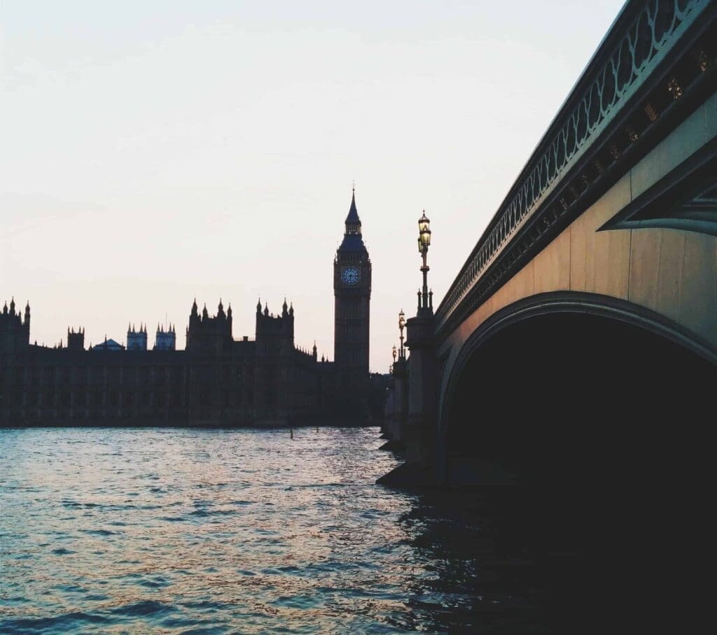 A view of the River Thames in London, with the iconic Big Ben clock tower and the Houses of Parliament on the left. Westminster Bridge spans the right side of the image, casting reflections in the water at dusk.