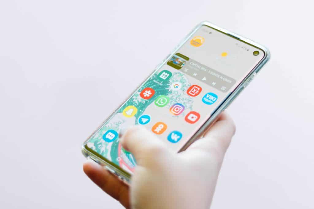 A person holds a smartphone with a clear case. The phone's screen displays various colorful app icons, including WhatsApp, Instagram, Snapchat, and Facebook Messenger, on a white and green background. The phone is held in the person's left hand.