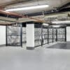 A modern, well-lit indoor bicycle storage area with metal 50mm x 50mm mesh security fencing and racks. The flooring is smooth and clean, with exposed pipes and lighting fixtures visible on the ceiling.