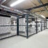 A large, well-lit bike storage area with metal mesh security fencing enclosing multiple bike racks of stored items. The ceiling features numerous pipes and ductwork, and the floor is clean and empty with a smooth, grey finish.