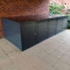 A row of four black metal E-Bike storage lockers is placed along a brick wall in an outdoor area with a tiled floor. The weatherproof charging points ensure convenience under the stairs, surrounded by greenery.
