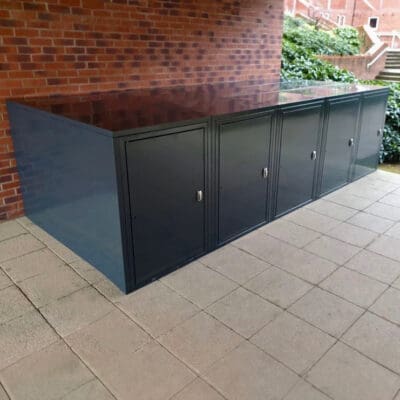 A row of four black metal E-Bike storage lockers is placed along a brick wall in an outdoor area with a tiled floor. The weatherproof charging points ensure convenience under the stairs, surrounded by greenery.