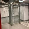 A secure storage area with metal wire fencing and a closed gate. The walls are white, and the floor is concrete. There are metal support beams and pipes visible overhead. The area appears to be part of a larger facility, possibly a warehouse or storage room.
