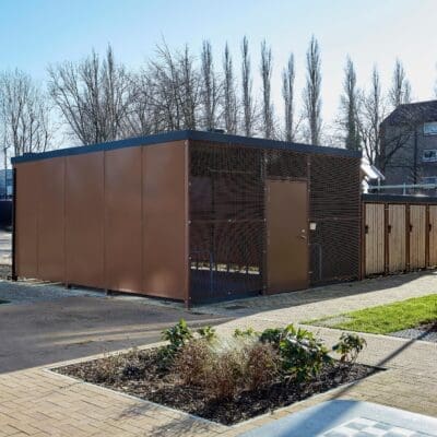 A large brown Carlton Shelter bike enclosure stands prominently beside a brick walkway, inviting visitors to explore its surroundings.