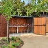 A modern outdoor bike parking area with a wooden fence and a covered metal rack with seven bicycles, surrounded by greenery and a brick wall.