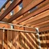 A wooden pergola stands in front of a brick building with large windows. The pergola casts shadows of its beams, creating a pattern on the wooden fence panels. The sky is clear, allowing for bright, natural lighting.