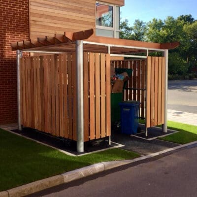 A small outdoor bin store with vertical wooden slats houses green and blue recycling and trash bins. The enclosure has a metal frame and a wooden slatted roof. It is situated between a paved road and a brick building, with a patch of grass on one side.