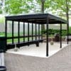 An outdoor pergola smoking shelter with a black metal frame and transparent panels on the sides, providing shelter over a seating area. The ground is paved with light and dark bricks, and there are trash bins and benches nearby, surrounded by green trees and bushes.