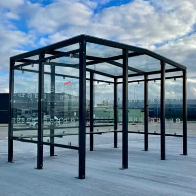 An image of the prestige smoking shelter, glass-walled, metal-framed smoking shelter stands on a paved surface under a partly cloudy sky. The surrounding area appears to be an industrial or commercial zone, with buildings and parked vehicles visible in the background.