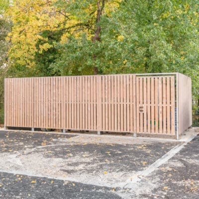 A wooden thurrock bin store enclosure is situated in a parking lot, with surrounding trees featuring green and yellow foliage. The enclosure has a gate and is designed to house waste and recycling bins. The ground is covered with asphalt, and some scattered leaves are visible.