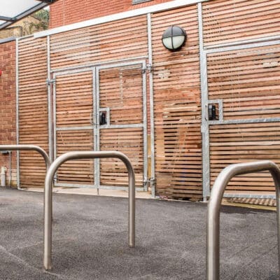An outdoor area featuring a metal bike rack with three sheffield stands on a paved surface. Behind the bike rack, there is a wooden fence with horizontal slats and a metal frame, along with a circular wall-mounted light fixture above a door.