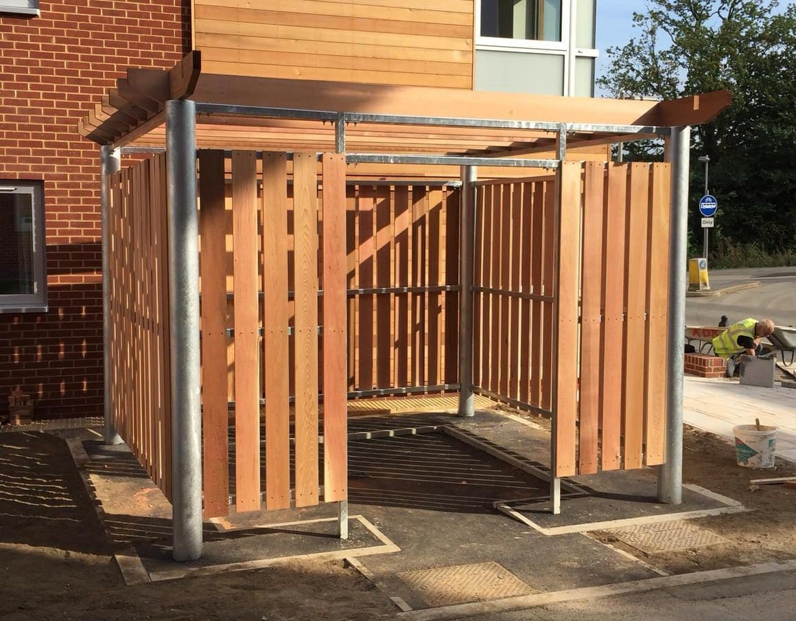 A wooden bin store with vertical slats stands on a concrete surface outside a red brick building. The structure is supported by metal poles and has a slatted roof, resembling a small enclosure or shelter.