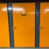 A row of bright orange storage lockers with dark gray frames is perfect for a Tenant Cycle Store, offering a high-security space. The middle locker has a metal sign displaying the number 13. All lockers have black handles and a smooth, shiny finish. The ceiling above has exposed red piping.