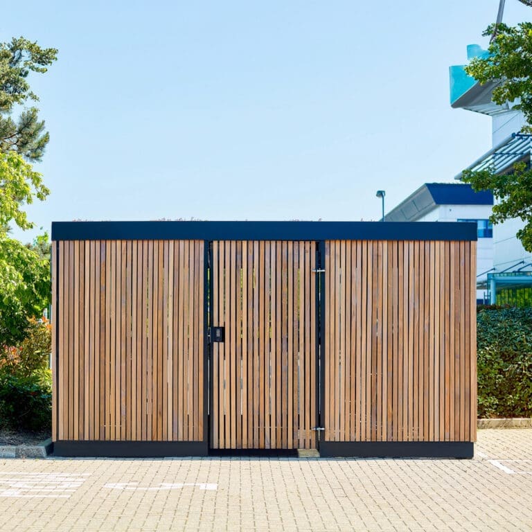 A modern wooden premium amazon eco enclosure with vertical slats stands on a paved surface surrounded by greenery on the left. The building in the background has a glass roof structure and a metal framework. The clear blue sky is visible overhead.