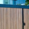 Modern wooden gate with vertical slats, a black metal frame, and a handle, set against a blurred background of a building with glass facades.