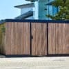A modern wooden bike storage facility with vertical slats, featuring a black bicycle symbol and double doors, located next to an urban building with a glass exterior.