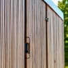 Vertical wooden slats form a wall with a sharp geometric metal handle on a door, highlighted by sunlight. a small security camera is mounted near the top of the frame.