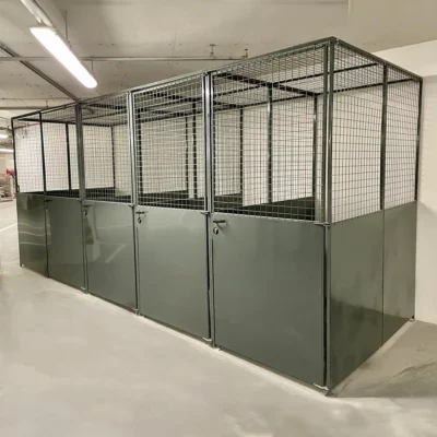 A row of secure, metal storage cages with solid lower panels and wire mesh upper sections, each with a lockable door, situated in a clean, well-lit indoor space. These mesh security cages are identical and aligned neatly side by side, ideal for the Battersea Tenant Cycle Stores in the new building development.