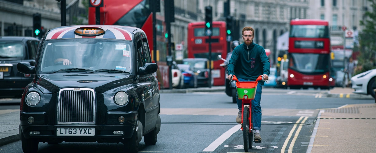 A person biking down a street in London on an electric bike and on his left there is a black cab.