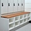 A white locker unit with keypad locks and a wooden bench surface sits over a series of open cubby holes. The cubbies provide extra storage space and are set against a white wall with a grey floor.
