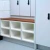 A modern room with white cabinets and lockers. Below the lockers, there is a wooden bench with storage compartments underneath. The lockers have digital locks, and the area is clean and well-organised, featuring a light grey floor and a white brick wall.