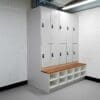 A modern locker room with white lockers, each equipped with a black lock. There are two rows of lockers stacked vertically and beneath them is a wooden bench. Under the bench are cubby compartments for storage. The room has white brick walls and grey flooring.
