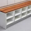 A white, multi-compartment storage bench with a wooden top. The bench has six open cubbies and is placed against a wall with additional cabinetry above it. The design is simple and modern, suitable for organizing and seating.
