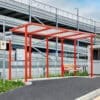 A modern bus stop with a bright red frame and a glass roof stands next to a fenced parking structure. The area is clean with a smooth asphalted surface, and a wooden bench is situated under the shelter. A bus stop sign is visible on the left.