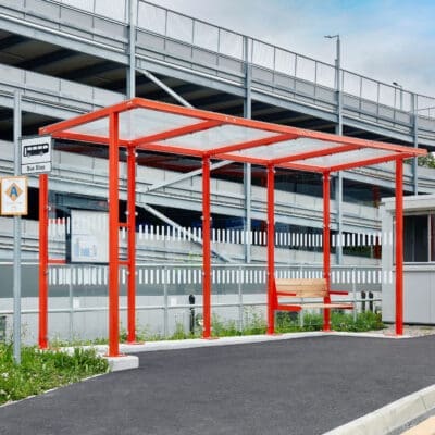 A modern bus stop with a bright red frame and a glass roof stands next to a fenced parking structure. The area is clean with a smooth asphalted surface, and a wooden bench is situated under the shelter. A bus stop sign is visible on the left.