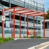 A red-framed bus stop with a transparent roof and side panels, situated along a paved road with a high-rise parking structure in the background. The bus stop has a bench and is surrounded by some greenery. Signs indicate it is a designated bus stop area.