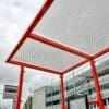 A bus stop with a red metal frame and a perforated metal roof. There is a bus stop sign on the left, and a modern building with glass windows in the background. The sky is cloudy.