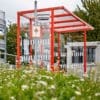 A modern red-framed bus stop with a glass shelter sits in front of a green area with wildflowers. There's a "Bus Stop" sign on top of the shelter and various plants and trees in the background on a cloudy day.