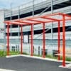 A bright red bus shelter stands on a black pavement before a multi-story parking garage. The shelter, which has a simple modern design and one small bench, is open on four sides and topped with a flat roof. A schedule or map is displayed on one of the side posts.