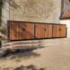 A row of bespoke cycle storage lockers with vertical slats is positioned along a stone wall. The lockers are secured with metal hinges and locks, and the ground is paved with a combination of concrete tiles and cobblestone edging. Trees are visible in the background.