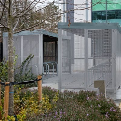 a bike rack and metal/mesh bike shelter behind a group of plants