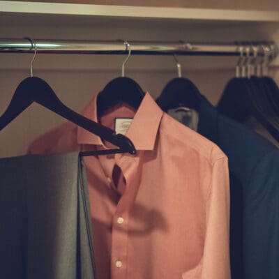 A neatly organised closet with several possessions hanging on black hangers. A peach-colored button-up shirt is prominently displayed, along with a gray pair of trousers draped over the hanger. Dark suits and other clothing items hang in the background, all benefiting from versatile storage solutions.