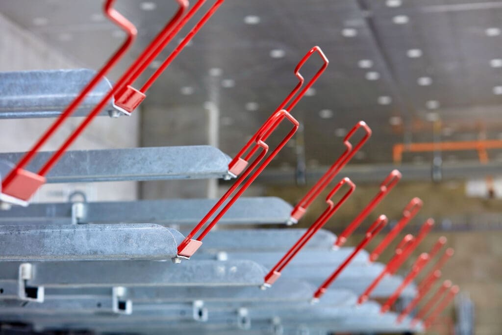 Close-up of red EasyGrip Two Tier Bike Rack handles aligned in a row, each attached to gray metal beams in an industrial setting. The background is blurred, showing ceiling lights and structural elements, emphasizing the orderly and symmetrical arrangement of the hooks and beams.