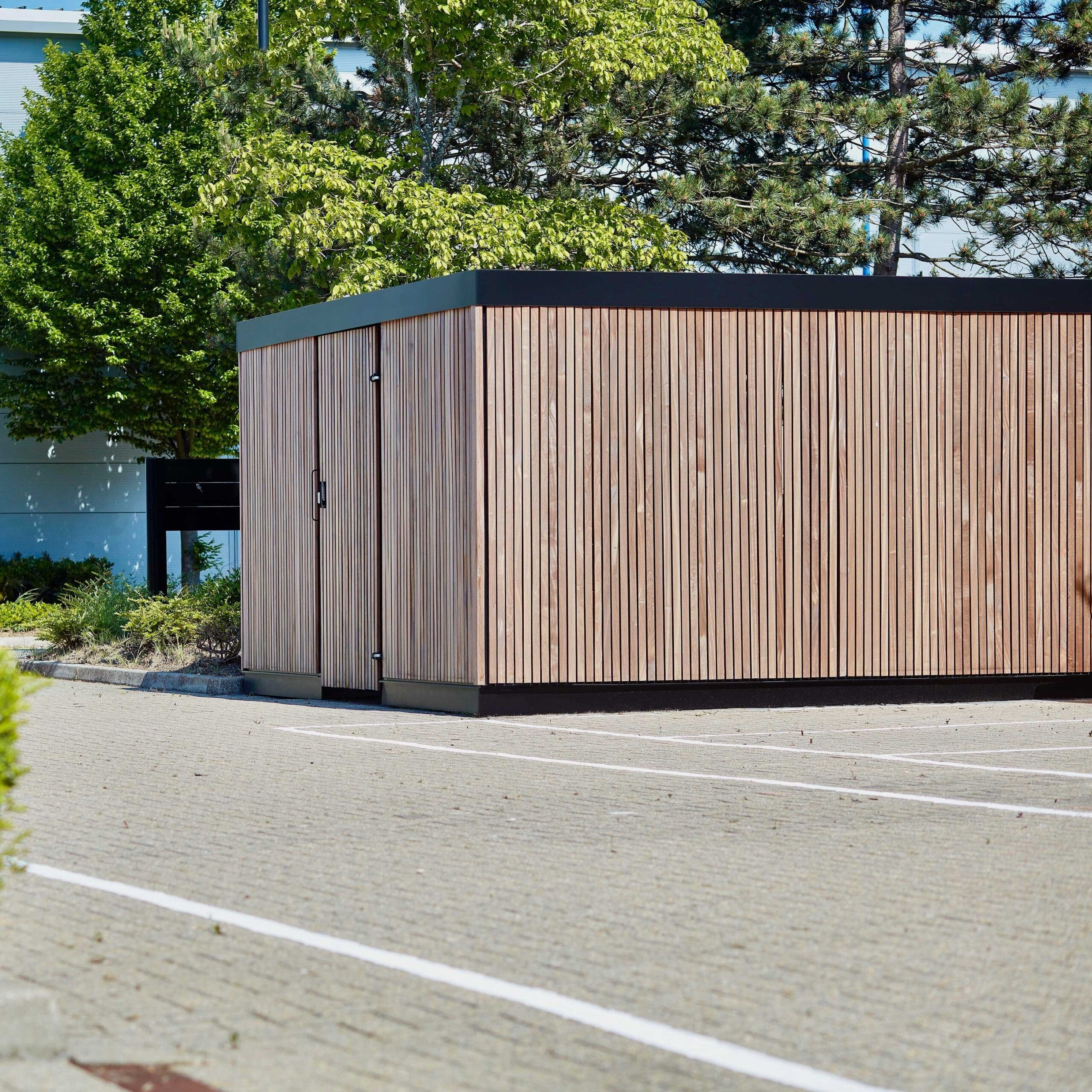 A Premium Amazon Eco Enclosure with vertical slats and a black roof sits in a paved, parking lot-like area. Trees with green leaves and some additional foliage are visible in the background. The weather appears to be clear and sunny.