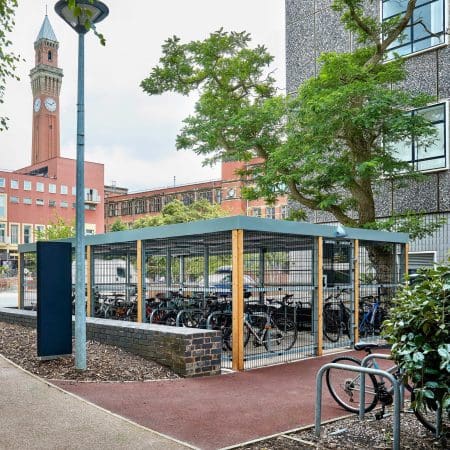 A fenced bicycle parking area is filled with bikes near a university campus. In the background, there's a tall clock tower and several university buildings. The area is surrounded by trees and a lamppost is located near the bike parking.
