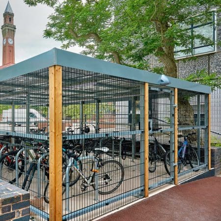 40 space security cycle enclosure bicycle parking area with a metal fence encloses several parked bicycles. Trees and a building with large windows are visible in the background, along with a tall clock tower. The area is paved with a dark surface.