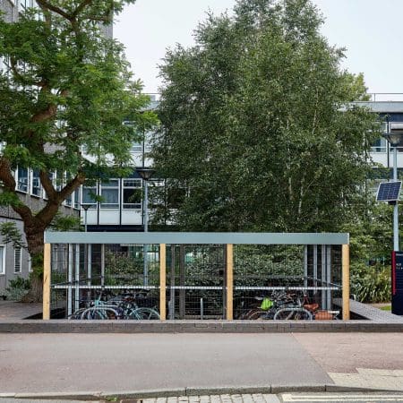 A bike shelter with a light blue roof and transparent panels, partially obscuring multiple parked bicycles underneath. The shelter is surrounded by trees and positioned in front of a modern building with many windows. A solar panel is visible on the right side.