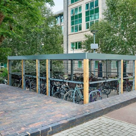 A bike storage area filled with bicycles is surrounded by a metal fence with a gray roof and wooden support posts. The storage is adjacent to a brick pathway and located near a multi-story building with large windows and surrounded by trees.