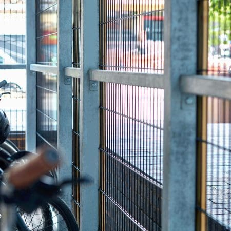 A close-up of a bike rack area enclosed by metal fencing. In the foreground, parts of bikes are visible including handlebars and wheel frames. Sunlight filters through the bars, casting shadows on the ground. A few vehicles can be seen in the blurred background.