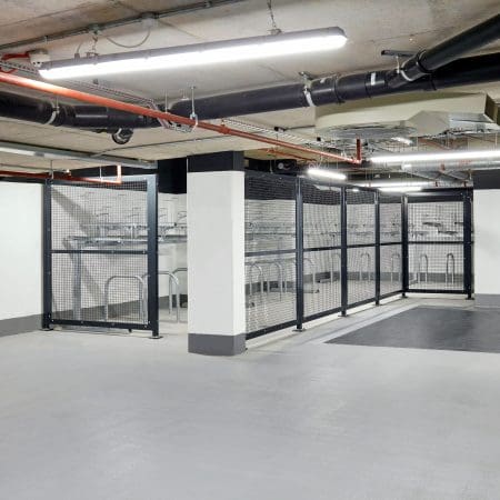 A modern, well-lit indoor bicycle storage area with metal 50mm x 50mm mesh security fencing and racks. The flooring is smooth and clean, with exposed pipes and lighting fixtures visible on the ceiling.