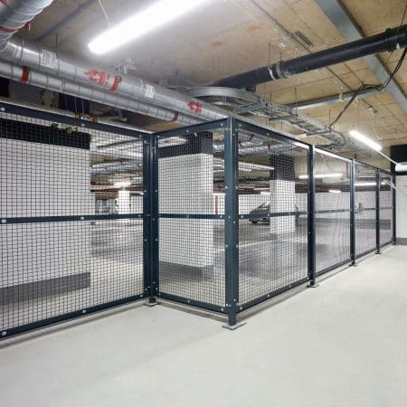 A large, well-lit bike storage area with metal mesh security fencing enclosing multiple bike racks of stored items. The ceiling features numerous pipes and ductwork, and the floor is clean and empty with a smooth, grey finish.