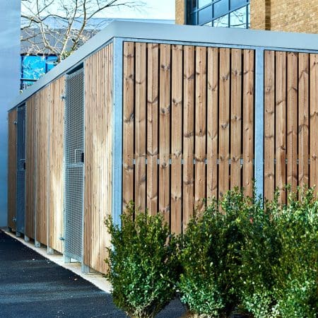 wooden cycle shelter with green shrubbery in-front and mesh swing door