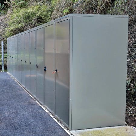Image of a row of large, grey vertical bike lockers situated outdoors along a paved path. The lockers are positioned against a retaining wall covered in greenery, extending into the background.