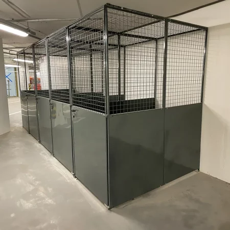 A spacious warehouse in the heart of a new building development features gray metal mesh security cages lined up in rows. Each cage has mesh walls and solid lower panels. The floor is clean, and the area is well-lit with overhead fluorescent lights.