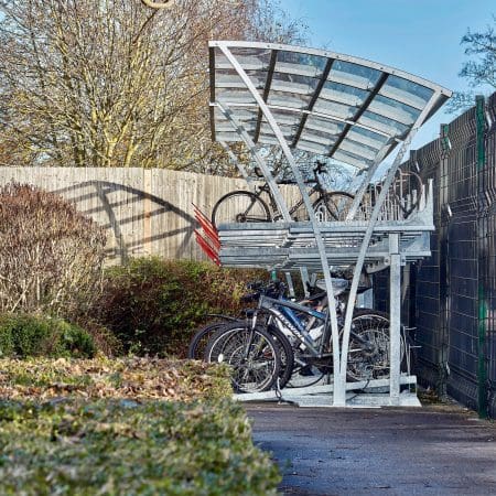 A metal, two-tier bike rack within a Chelsea Cycle Shelter holding multiple bicycles, located next to a fence and shrubbery. Some bikes are parked on the upper rack while others are on the lower level. Trees are visible in the background.