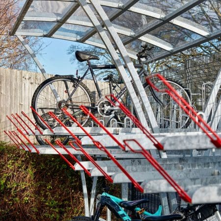 A modern, two tier bike rack holds several bicycles in an outdoor area. The structure is made of metal with a transparent roof. One black bike is parked on the upper level, and a blue and black bike is on the lower level. Trees and a wooden fence are in the background.